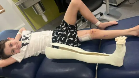 Laura Ducker/PA Wire Alfie is lying on a hospital treatment bed next to a large leg cast. He has his hands behind his head and he is smiling at the camera.