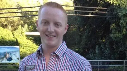 Kevin Bell, a man with short, ginger hair, stands on a train platform on a sunny day. He is smiling and wearing an open-necked checked shirt. There are tall trees in the background.
