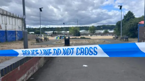 Kate Bradbrook/BBC Blue-and-white police tape stretches across a pathway in the foreground, blocking access to an open area of Kingsway Recreation Ground. Several benches and lamp posts are visible beyond the cordon, with a police vehicle parked in the distance. A large building stands to the left and trees line the background beneath a cloudy sky.