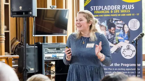 Ben Gregory-Ring/Tring Park School Josie Rylance, standing a room with a sign behind her and screens and equipment to her right. She is mid-clap and is smiling. She is wearing a blue and black checked dress, and has long blonde hair. 