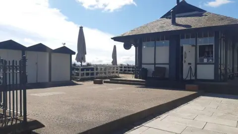 LDRS The patio and side entrance of the former tram shelter, showing three black beach huts with white painted doors.