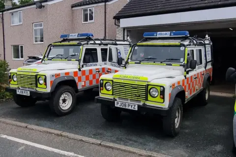 Penrith Mountain Rescue Team Two mountain rescue vehicles are parked on a residential driveway in front of an open garage. They have fluorescent yellow/orange patterns on them and blue lights on top.