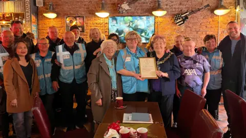 Gemma Sherlock/BBC A group of 20 people stand and smile at the camera in a cafe, holding a framed award 