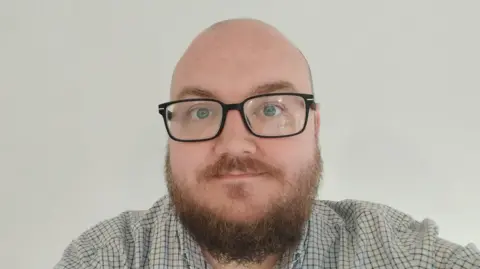 A selfie of a man wearing glasses and a shirt while standing in front of a white wall. 