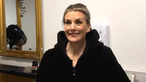 Colleen Mullan stands in her hair salon, she has blonde hair puled back and is wearing a black hoodie. Behind her to the left of the image is a gold-framed mirror in which a large black blow-dryer can be seen.