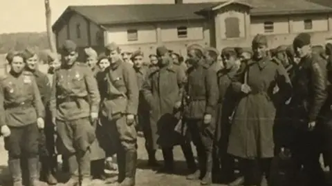 John Lloyd A black and white photo from spring 1945 at Wismar, Germany. It shows a large group of Russian soldiers and in the middle can be seen Jack Lloyd in his British Army beret. Behind them is a low building. 