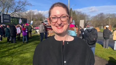 Katie Sang wearing pink coloured glasses and a black top. She is smiling at the camera and is standing in front of a crowd of people on a grass verge.