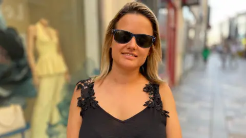 Julie Hristov has shoulder length blonde hair, is wearing sunglasses and a black summer top. She is standing in the high street.