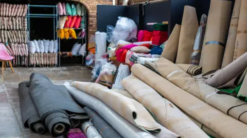 KDM Photography A pile of rolled up carpets on stone tiles. In the background there are shelves with cushions