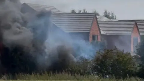 Black smoke in front of a new housing development. The smoke is spreading across a nearby field. 