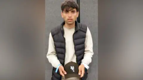A young man stands in front of a camera. He is holding a cap in his hands. 
