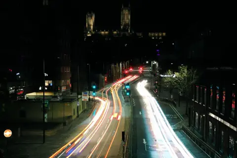 Graham Halliday A photo taken at night showing light trails from car lights snaking along a road towards a large cathedral on a hill, which is partially lit by spotlights.