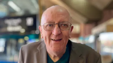 Martin Giles/BBC Steve Hewitt is wearing a light brown jacket over a dark green top. He is also wearing glasses.