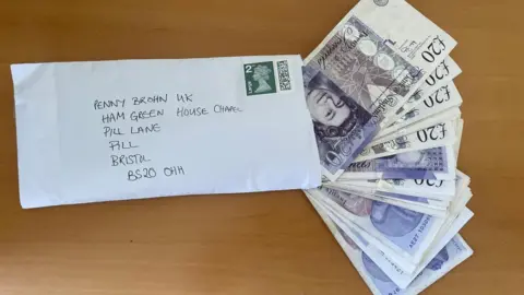 Penny Brohn UK A white, stamped, handwritten envelope with wads of old-style twenty pound notes fanned out of it from an opening on its right edge edge