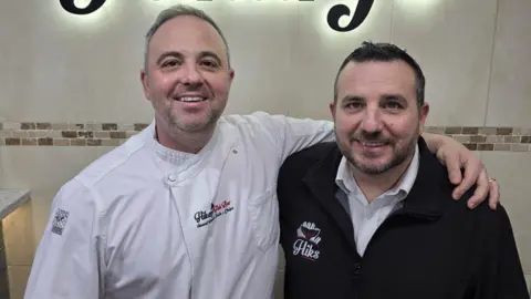 Two men are smiling as they stand next to each other in a fish and chip shop. They are both wearing branded tops, with Ouday on the left wearing a white chef's outfit with 'Hikary's Fish Bar' on it. Emir is wearing a black fleeced top with Hiks printed on it.