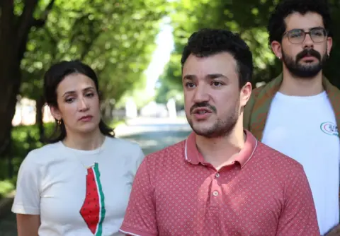 Mahmoud Khalil wears a pink shirt outdoors and is flanked by two people in white shirts. 