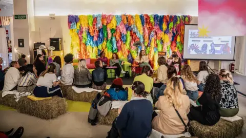 Sistaland Women sitting on the floor listening to three women sat on armchairs. There is a rainbow coloured design begin the speakers. 