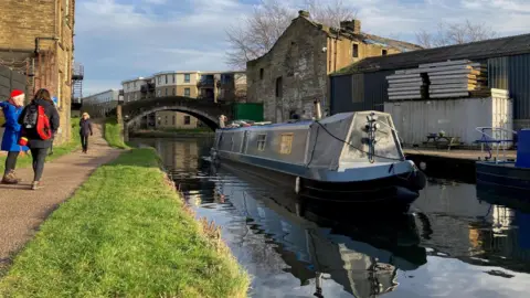 Charles Heslett/BBC A grey narrowboat cruising down a canal with people walking on a towpath