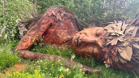 A larger than lifesize statue, carved out of wood, of a woman with a crown of leaves. She lies on her side in the middle of shrubs and trees, with her eyes closed as though asleep. There are smaller plants with white and yellow flowers in the foreground.