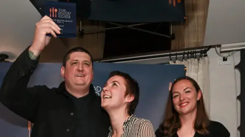 Getty Images Chef Gareth Ward stands on a small stage in front of a “The Good Food Guide” backdrop. He raises an award plaque above his head while smiling, his guest, a woman, looks up at it, smiling, and a woman on the right smiles while holding a microphone and a card.
