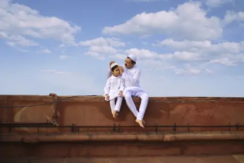 Jobit George / National Geographic A father and son sitting in white traditional attire with blue sky behind