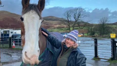 Cumbrian Heavy Horses riding centre owner Annie Rose standing in the yard of the riding centre with one of her horses. The horse is a large, heavy horse breed and is brown with a white face. Rose is resting her hand on the horse's shoulder.