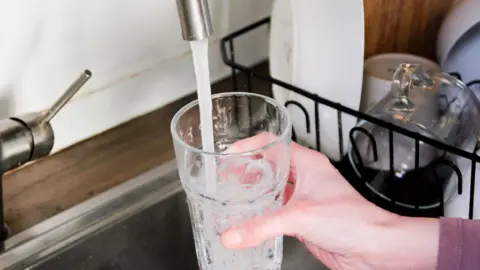 A hand filling a glass of water up from a tap, with dishes in the background