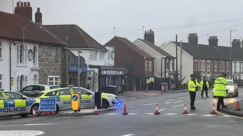 Angie George/BBC A road, with a number of houses and shops on it, with a number of police officers in high-vis jackets in it. There are two police vehicles and traffic cones in the street. 