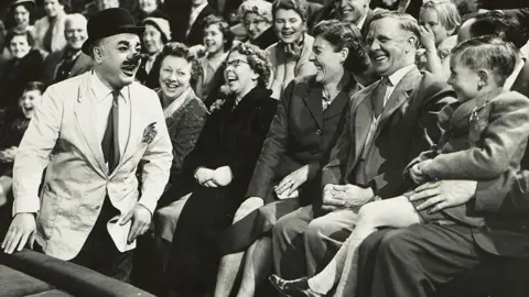 Barnet Saidman Black and white image of Charlie Cairoli ringside by a crowd of laughing people looking towards a laughing child. He is wearing a bowler hat, white jacket, shirt and tie and has a big clown nose, large eyebrows, a moustache and dark lips