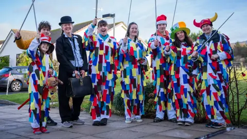 Brook Wassall A group of people wearing white costumes decorated with colourful strips hold prop swords above their heads. There is also one lady wearing a bowler hat, dark suit and holding a suitcase. They are all smiling and posing for the camera and are standing outside on a pavement.