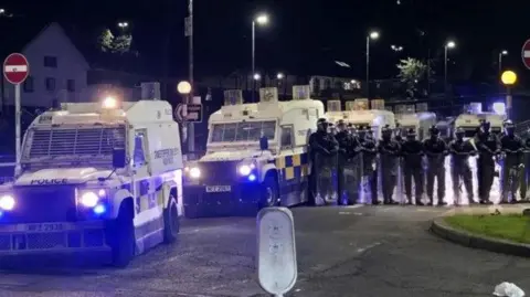 A number of white police Land Rovers with more than a dozen riot police in black and wearing helmets and carrying transparent shields.