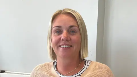 A head and shoulders shot of Claire King, who has a blonde bob haircut. She's smiling at the camera. She is wearing a peach coloured cardigan over a black and white stripped top and a silver necklace.