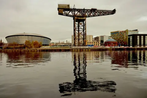 Graham Christie A large decommissioned crane stands reflected in the River Clyde. Buildings are visible in the background including the oval-shaped OVO Hydro concert and event venue