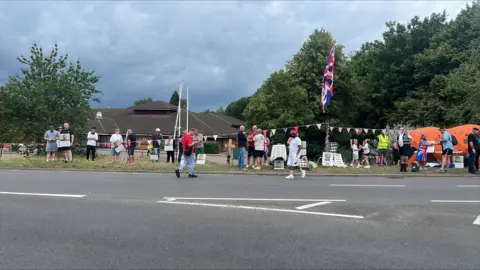 BBC/Richard Knights A group of people, mostly standing, by a roadside. There is bunting tied up and a union jack flying.