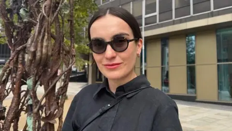 Polish artist Alicja Biala, wearing a black shirt dress and round sunglasses. She has dark brown hair which is tied back in a tight bun. 
