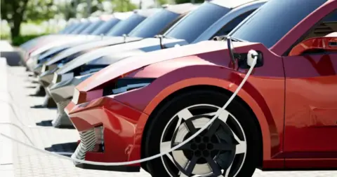 A row of cars parked at electric charging points. The nearest car is red.