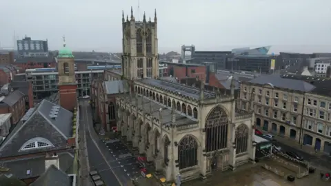 An aerial view of Hull Minster taken using a drone. The gothic building with a large arched entrance stands in a square surrounded by other modern buildings. The Hull tidal barrier and a pyramid-shaped building are visible in the background.