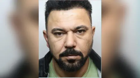 Home Office Mugshot of Mukhlis Jamal Hamadamin. He has black hair and a trimmed beard. He is photographed wearing a green t-shirt and black jacket.