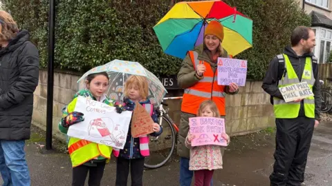 Becky Carlyle Children and adults stand on the pavement in protest holding signs. Two of the children are huddled under an umbrella. One sign reads: "No cars on corners." Another says: "I run on the path." Another says: "Safe pavements for people." The fourth sign says: "Can't park there, mate."