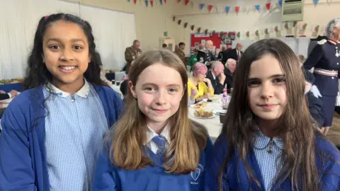 Three young girls wearing blue uniforms. They are smiling at the camera while standing in a classroom.