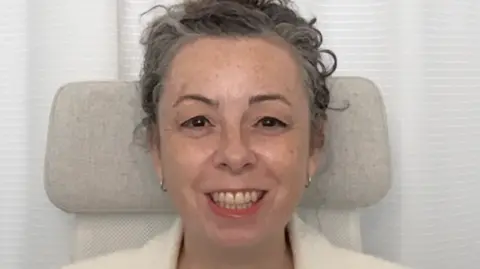 University of Worcester A woman with short grey hair smiles as she sits on a grey chair.