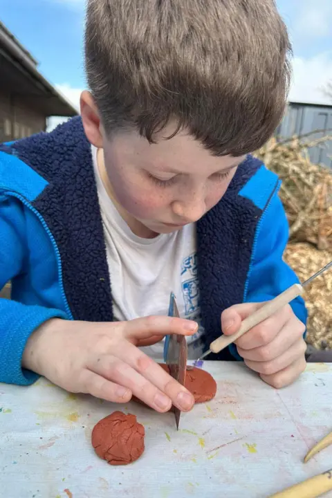 Necessary support group A young boy with brown hair sat at a table holding model clay. He had his head down as he works on the craft. He is wearing a white t shirt and blue hoodie and behind him are stacks of hay bails.
