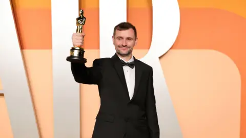 Rhys Salcombe holding his Oscar award up at the ceremony. He is wearing a black tuxedo