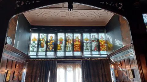 National Trust Stained glass windows at Wightwick Manor. There are nine long rectangle-shaped windows with figures on surrounded by dark brown wood. 