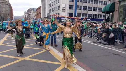 Dancers wearing golden, blue and black frocks hold sticks in the air as they dance in the street. Along the side of the route are people queued up, with photographers taking pictures.