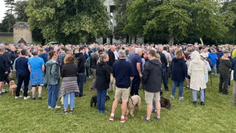 A crowd of people gather on the Downs near the water tower - they mostly wear waterproof coats and there are some dogs amongst them