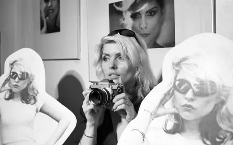 Martyn Goddard Debbie Harry holds a camera while surrounded by cardboard cutouts and portraits of herself in a black-and-white photo.