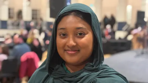 A picture of Aishah wearing a green hijab, smiling, and looking into the camera.