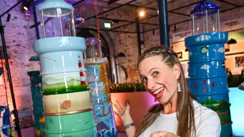 A woman with long brown hair smiling at the camera. She is standing in front of three lighthouse sculptures which are all brightly painted and lit