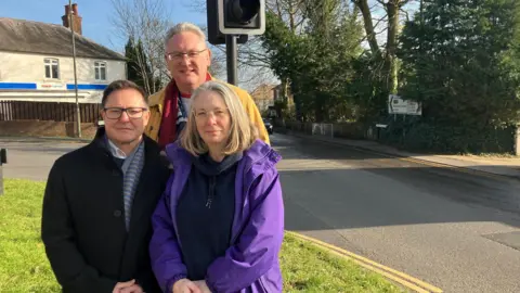 Councillor Catherine Powell, with residents Elliot Russo and Simon Cross standing by the traffic lights on Upper Hale Road. In the background is a branch of Tescos and signs directing cars where to go.